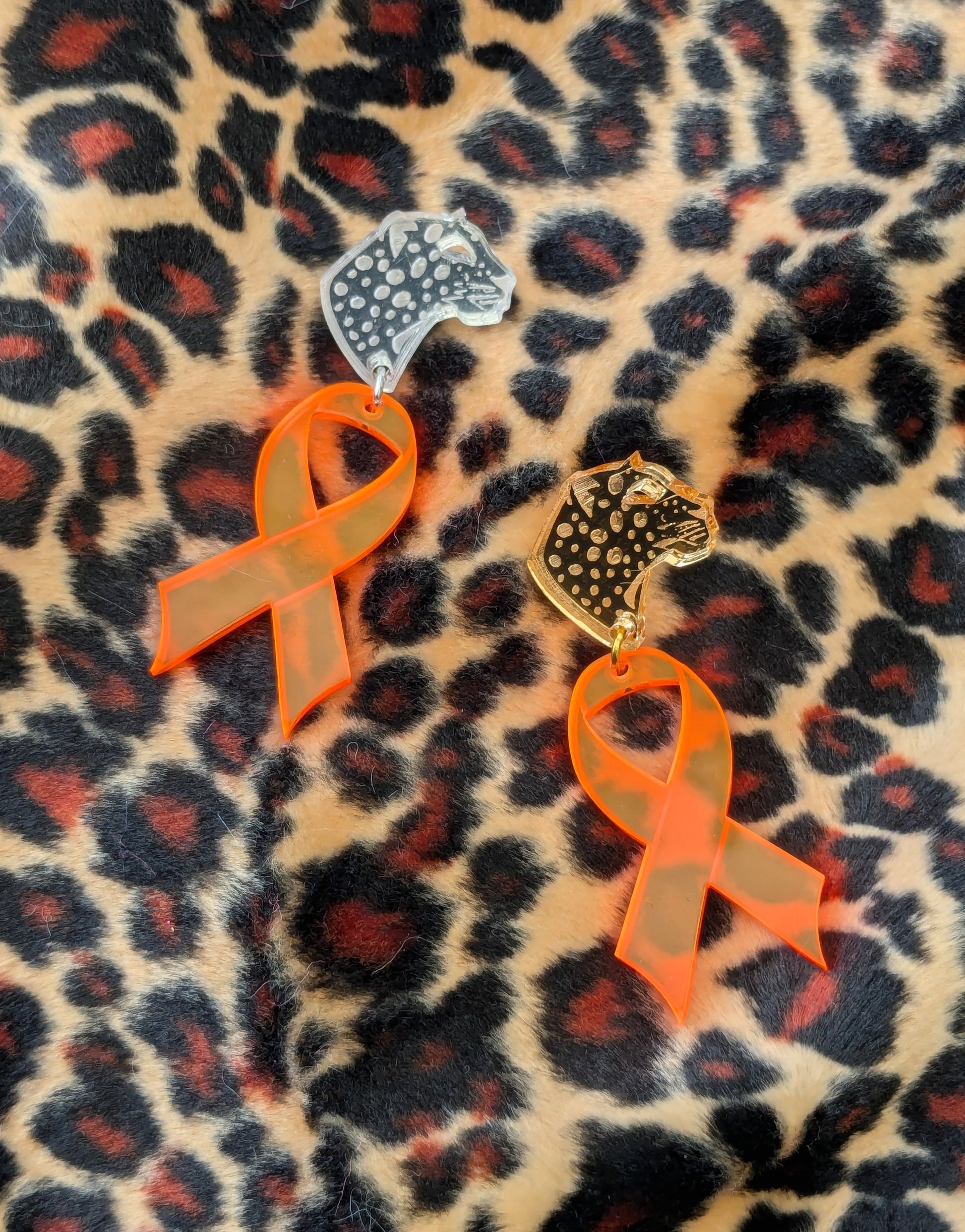 Two leopard head pins, one gold and one silver, both with orange acrylic ribbon attached laying on a velvety leopard print background