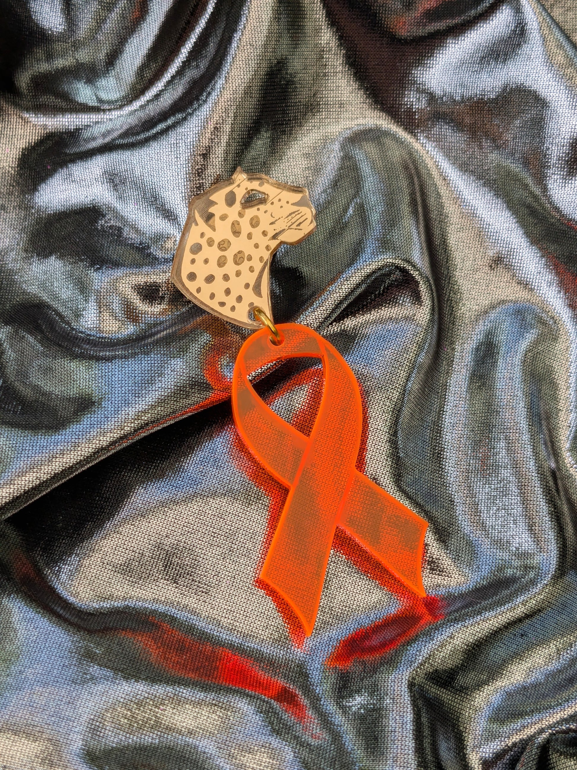 Gold leopard head pin, with orange acrylic ribbon attached on a flowing metallic silver fabric background