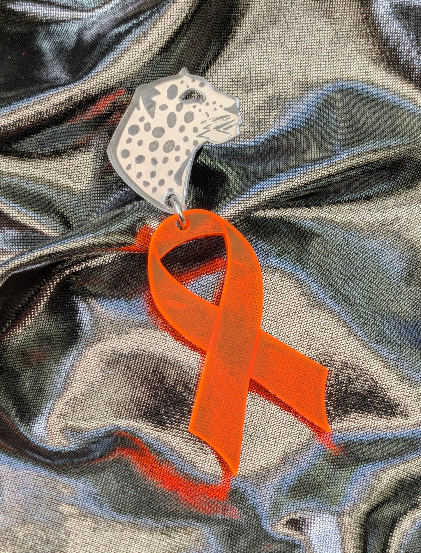 Silver leopard head pin, with orange acrylic ribbon attached on a flowing metallic silver fabric background