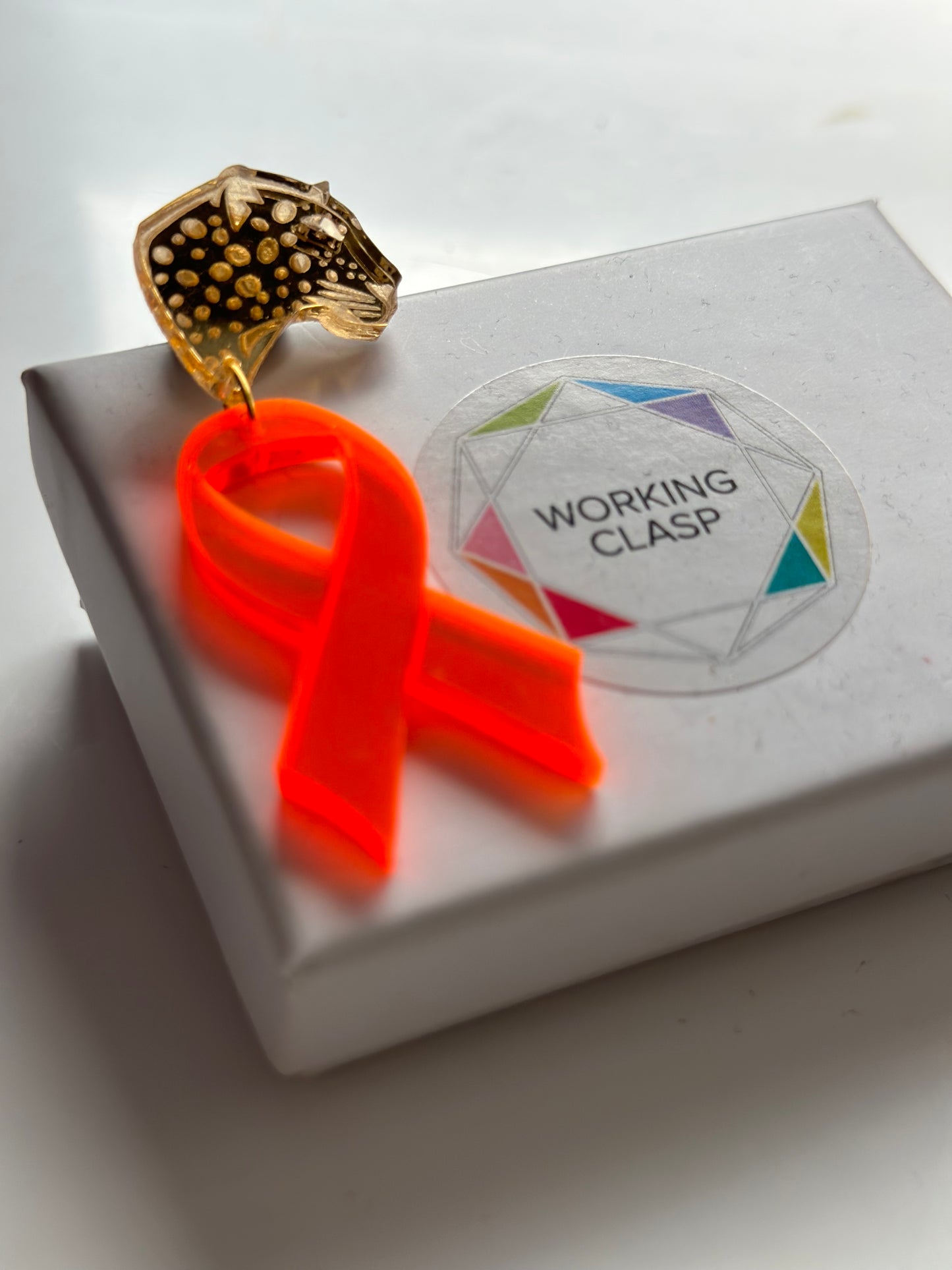 Gold acrylic pin of a leopards head attached to a bright orange acrylic ribbon sitting on top of a white jewellery box with the logo for jewellery brand working clasp